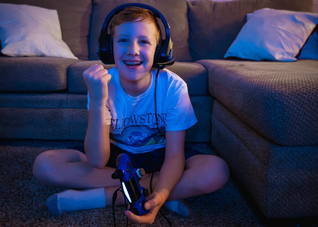boy smiling with braces on his teeth wearing gaming headphones and while holding a game controller sitting criss cross applesauce in the floor in front of a couch playing a video game with safe parental controls