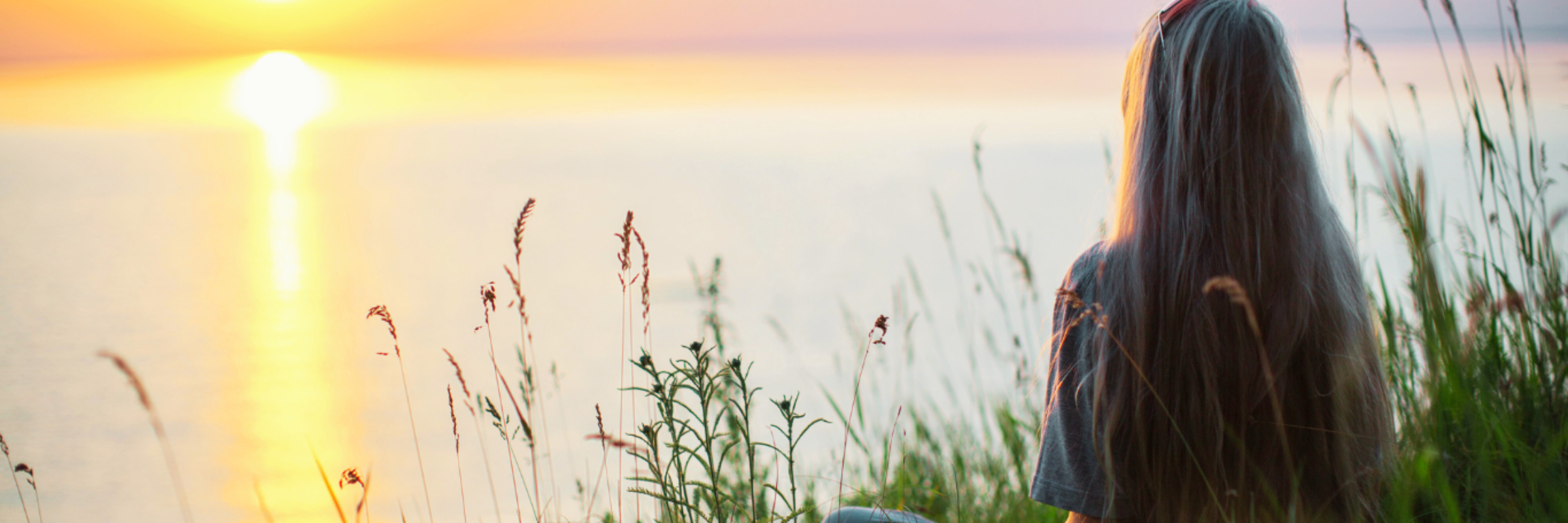 woman sitting Indian style in tall grass staring peacefully at the sun setting over a body of water