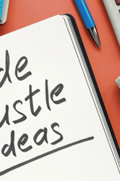 the words side hustle ideas written in black marker on a notebook lying on a desk. There is also rolled up cash, a pen, and a calculator