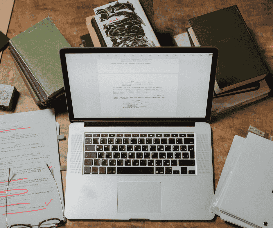 an open MacBook on a desk with paper, glasses, and stacks of books